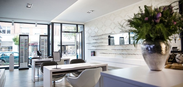 Modernes, helles Optikergeschäft mit Brillenregalen, Beratungstischen und Blumendekoration.